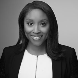 Black and white headshot of a dark skinned woman with shoulder length hair smiling at the camera with a black blazer on and a white dress shirt underneath that zips in the middle.