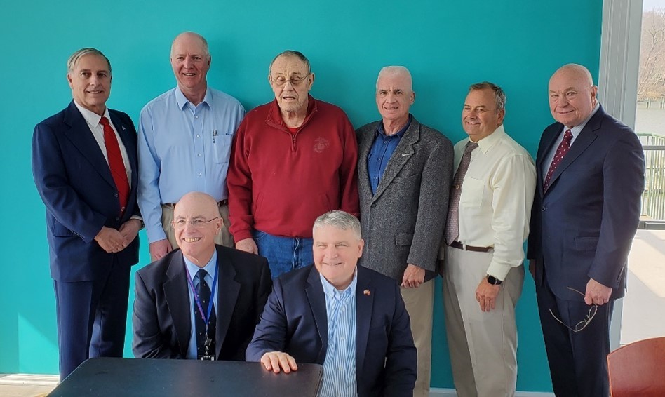 A group of eight men pose for a photo in front of a teal wall.