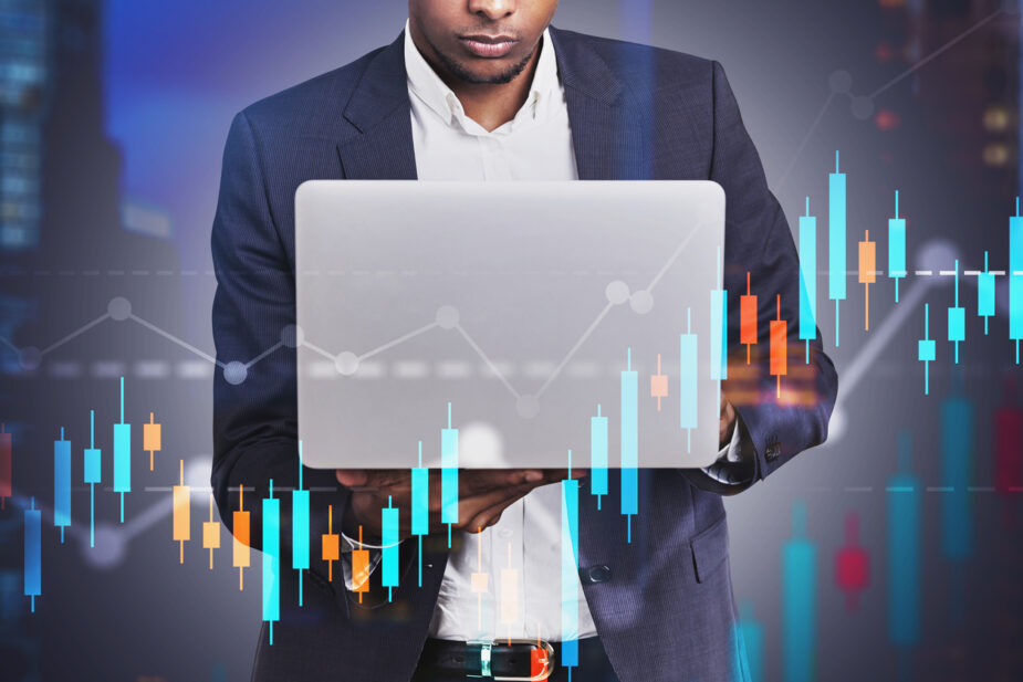 Serious young African American businessman working with laptop in blurred night city with double exposure of graphs. Concept of stock market.