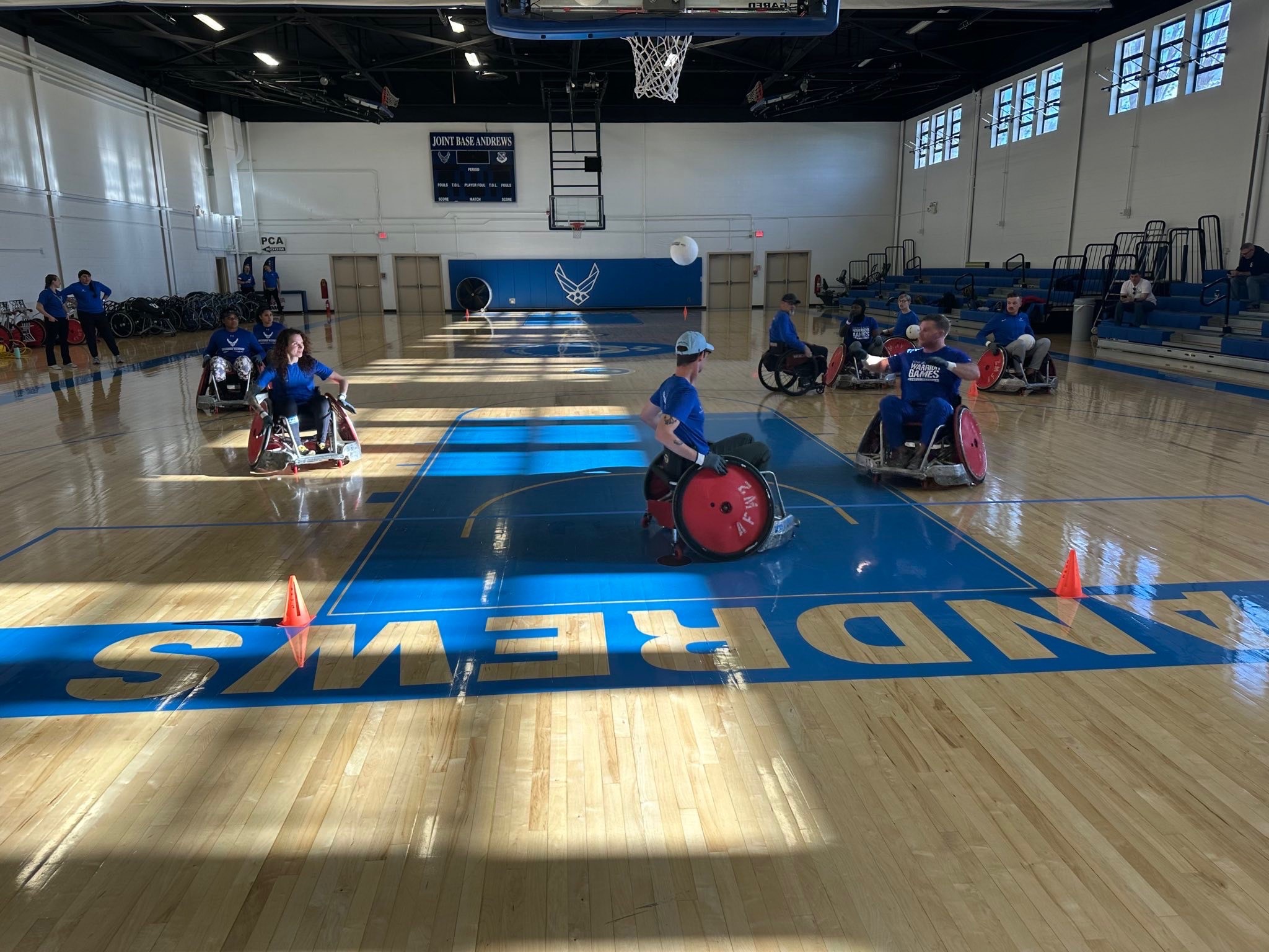 Several individuals in wheelchairs are participating in a sport on a basketball court marked with blue paint and orange cones. A basketball hoop is visible in the background.