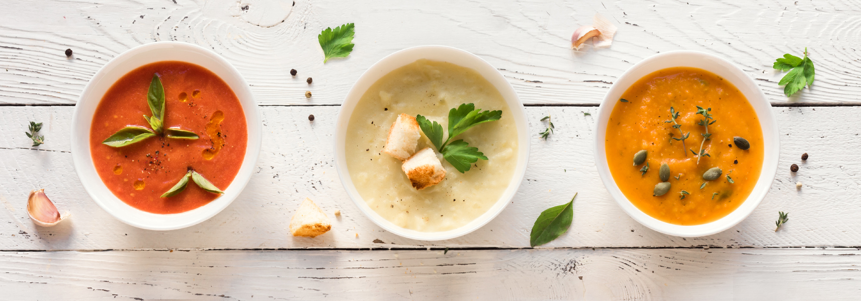 Three bowls of soup, each a different color (red, cream, and orange), are arranged on a white wooden surface with scattered herbs, spices, garlic cloves, and croutons.