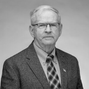 Black and white headshot of an older Caucasian male with white hair combed to the side and glasses. He is dressed in a suit with a plaid tie giving the camera a small grin.