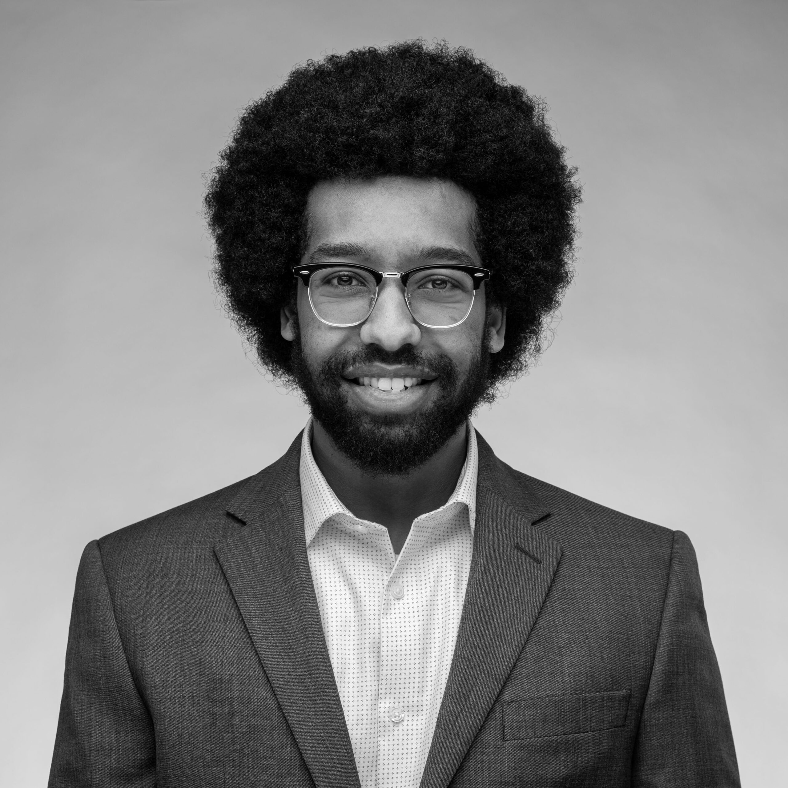 A black and white portrait of a smiling person with dark skin, an afro, glasses, and a beard, wearing a suit jacket against a gray background.