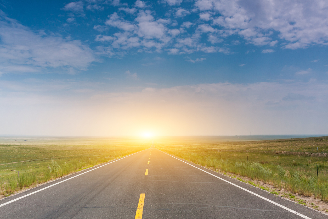Straight asphalt road stretching to the horizon with a bright sunburst at the vanishing point, under a blue sky with scattered clouds, and green fields on either side.