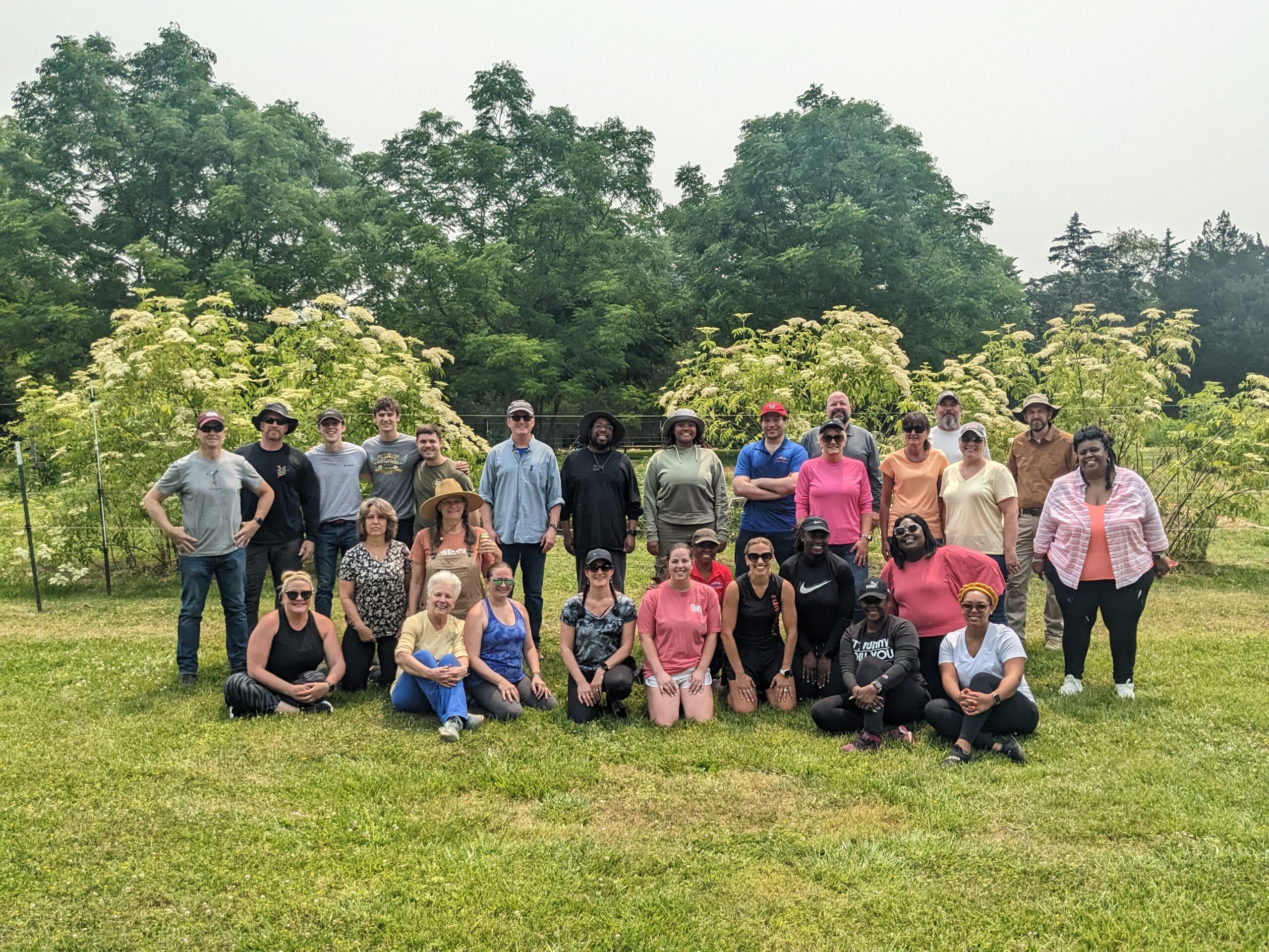 Group photo of approximately 15 people standing outdoors in front of trees and flowering bushes. The people are diverse in age and appearance, and are casually dressed. The sky is overcast.