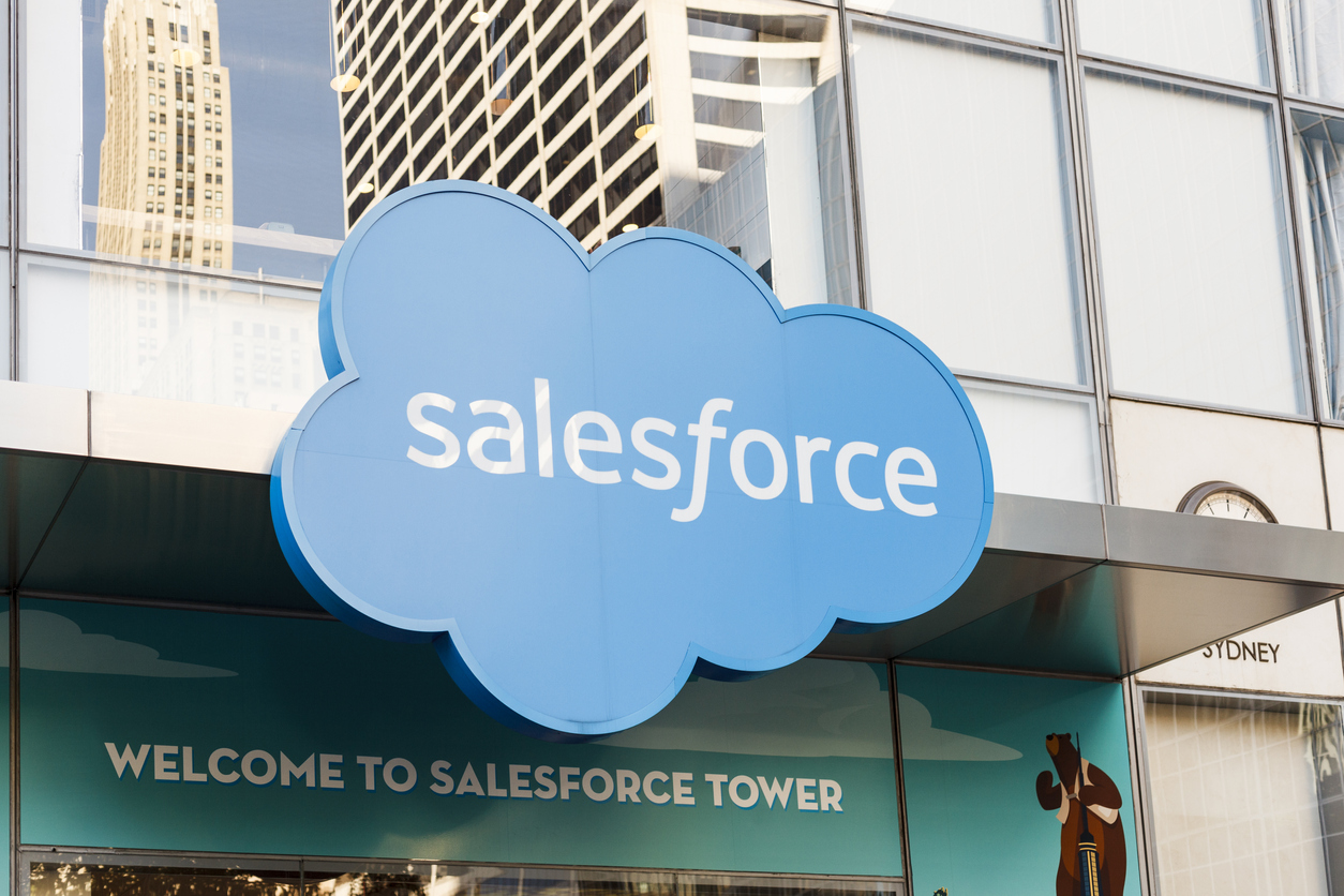 Salesforce logo, a light blue cloud shape with the word "salesforce" in white, affixed to the exterior of a modern building with glass and white paneling. Another tall building is reflected in the glass. The lower portion of the image is a solid light gray.