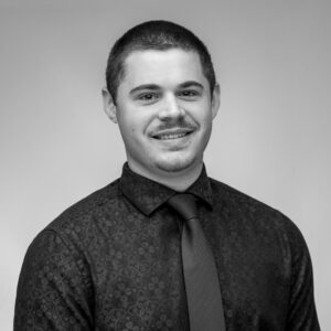 Black and white headshot of a young Caucasian man with a buzzcut, short mustache and beard, smiling at the camera in a dress shirt and tie.