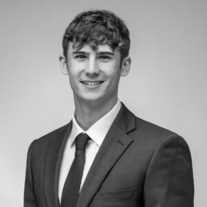 Black and white headshot of a young Caucasian man with curly hair smiling in a suit.