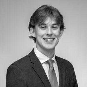 Black and white headshot of a young Caucasian man with longer hair smiling at the camera in a suit.