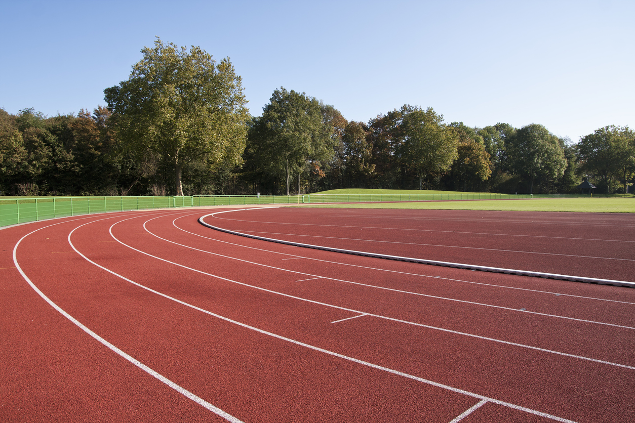 Empty red running track curving to the left, with white lane lines, green fence, trees, and blue sky.