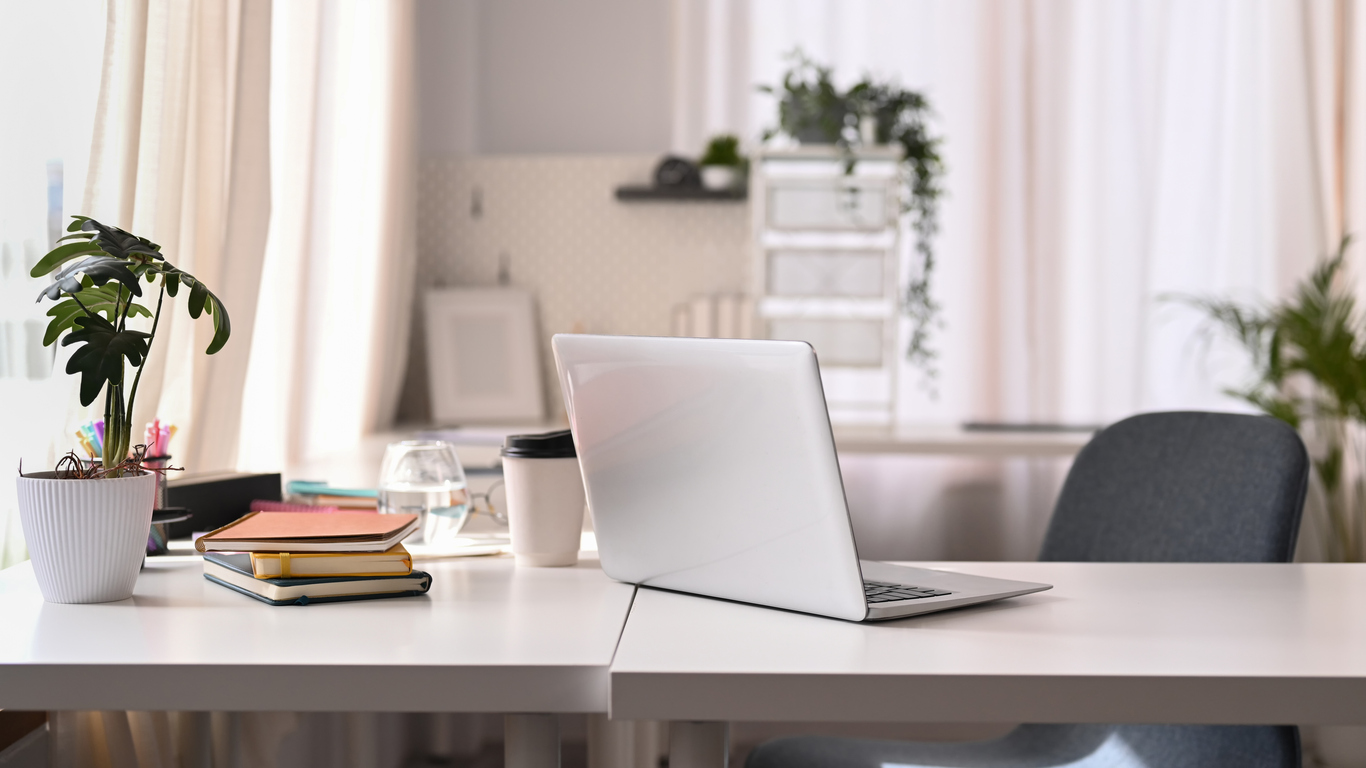 Workspace with laptop: White desk with a laptop, notebooks, coffee cup, and plant, with a blurred background of a window and curtains.