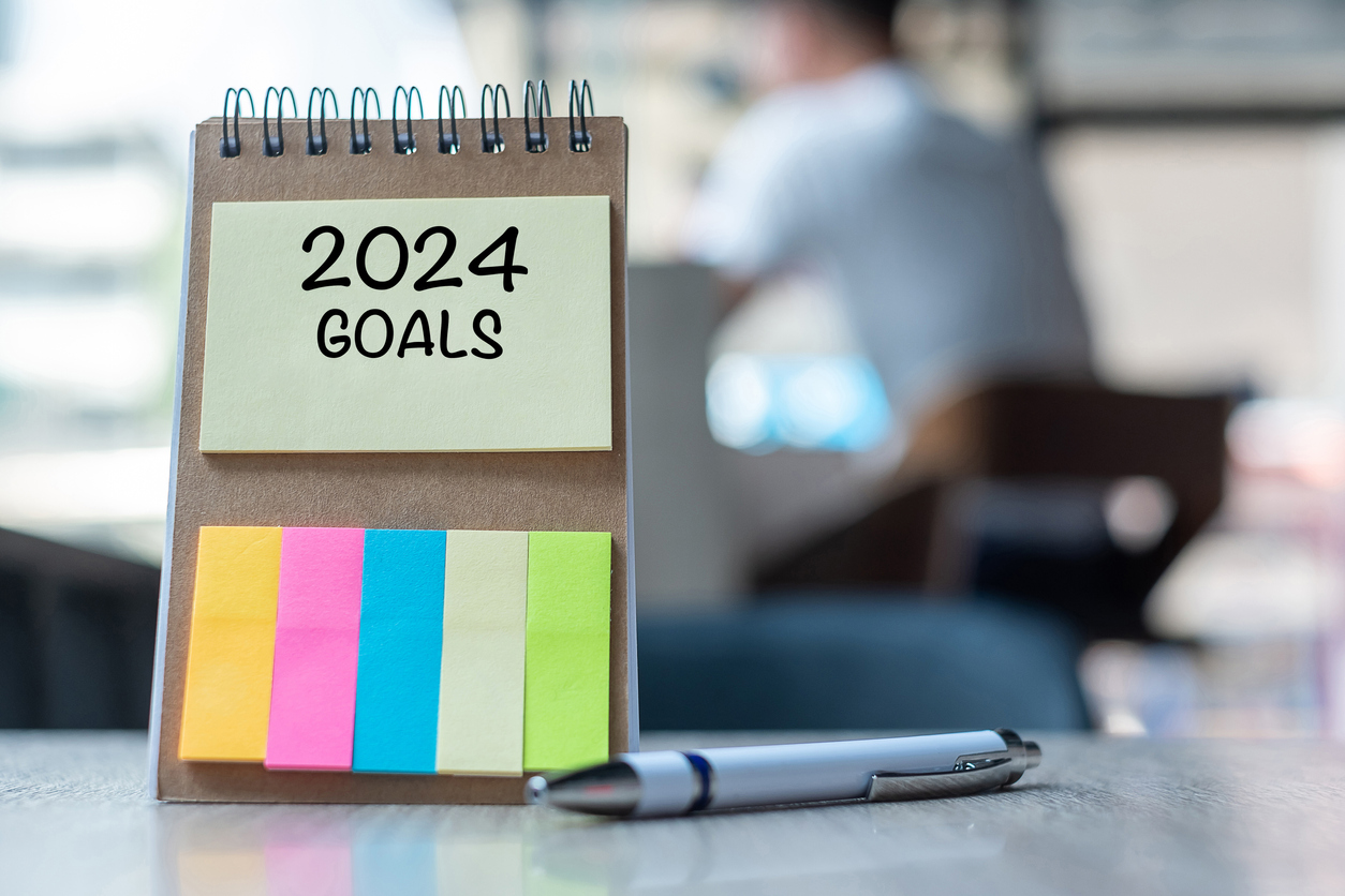2024 Goals concept: Notepad with "2024 GOALS" written on a yellow sticky note, colorful sticky tabs, a silver pen, and a blurred background of a person sitting in a chair.
