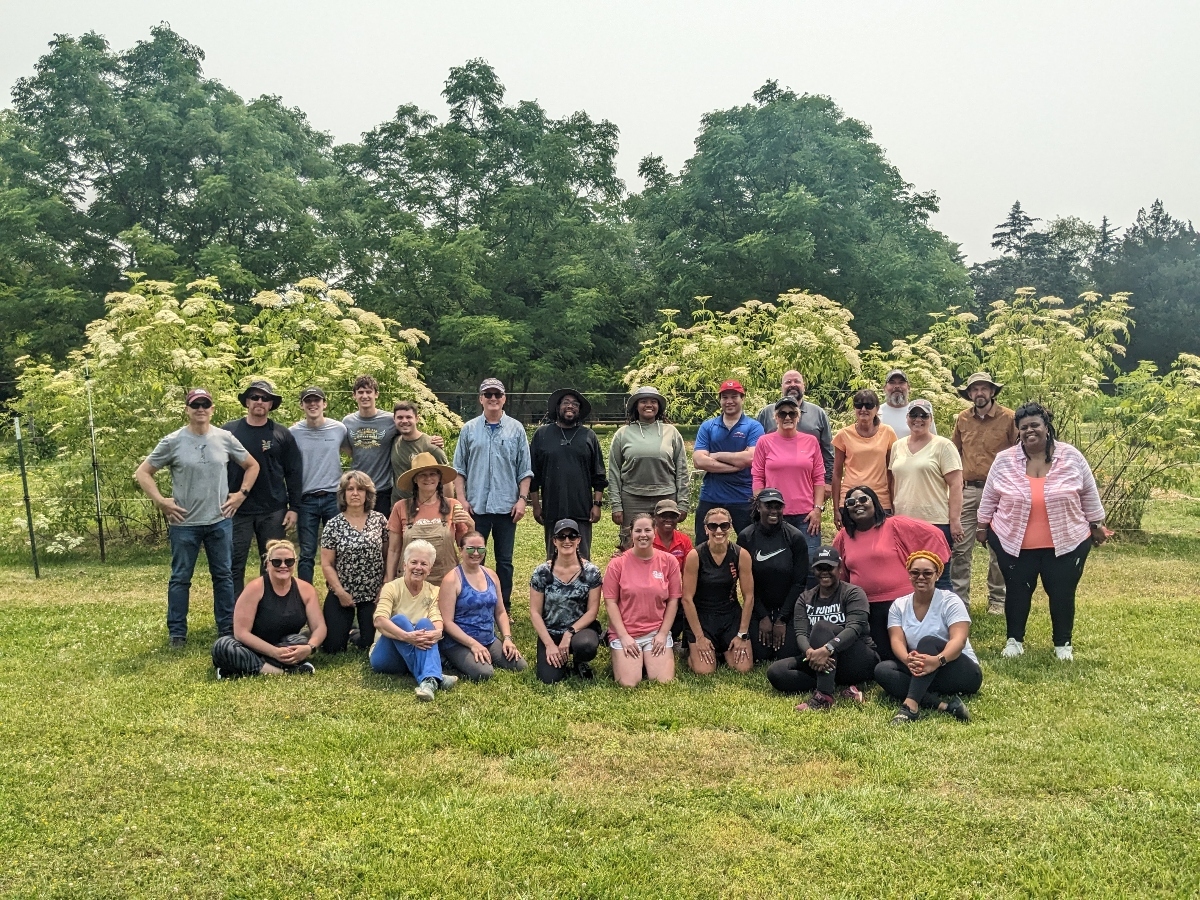 A large group of about 25-30 men and women of various ages are gathered on a grassy lawn, with some standing and others kneeling. They are all casually dressed and smiling at the camera, with trees and flowering bushes in the background.