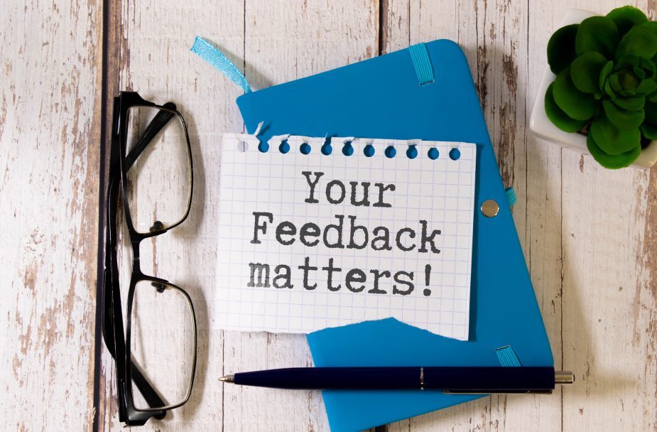 A note reading "Your Feedback Matters!" placed on a blue notebook, with glasses, a pen, and a small green succulent on a wooden desk.