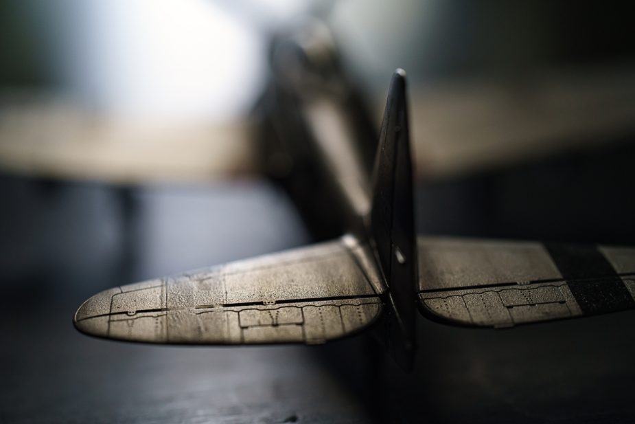 Close-up photograph of the tail section of a model Spitfire airplane, showing the horizontal stabilizers and vertical fin, with a blurred background.