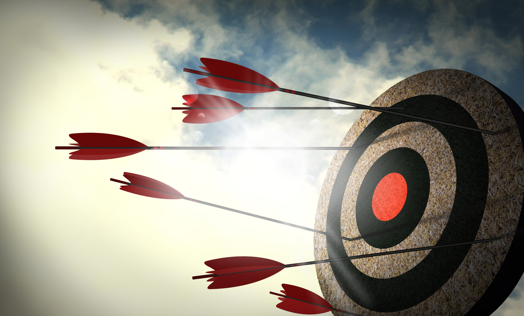 A circular target with concentric rings of brown and black, and a red bullseye, has five red arrows with black fletching and silver tips embedded in it. None of the arrows have hit the central red bullseye; they are scattered across the outer rings. The target is set against a backdrop of a partly cloudy sky with sunlight breaking through from the left.