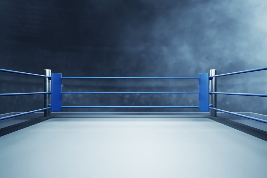 An empty boxing ring with a white canvas floor, blue corner posts, and blue ropes is shown against a dark, smoky background with a light source from the upper right.