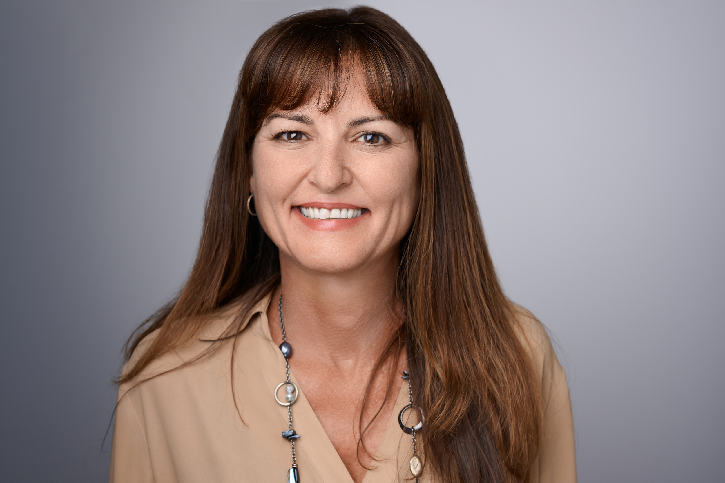 A smiling woman with long brown hair, brown eyes, and bangs looks directly at the camera. She is wearing a light tan blouse and a long beaded necklace. The background is a solid light gray.
