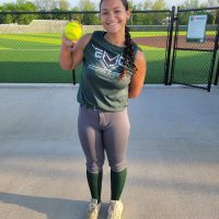 A young woman with dark hair in a braid smiles while holding a yellow softball on a paved surface next to a softball field.