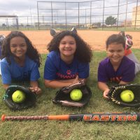 Three young softball players lie on the grass with their gloves and a bat, smiling at the camera.