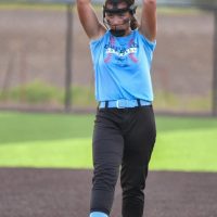 A softball player in a blue jersey and black pants prepares to pitch, wearing a face mask and standing on a dirt field.