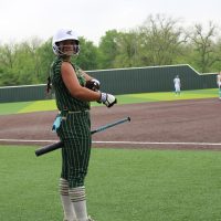 A softball player in a green and white pinstripe uniform stands on the field, holding a bat and adjusting her batting gloves, with other players in light blue and white uniforms in the background.