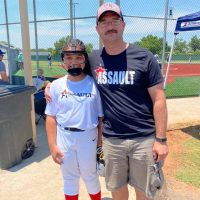 A young softball player in a helmet and mask poses with an adult man wearing a baseball cap and a t-shirt that says "ASSAULT" at a sunny outdoor baseball field.