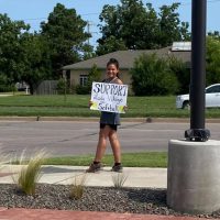 A young person stands on a sidewalk holding a sign that reads "SUPPORT Lady Vikings Softball."