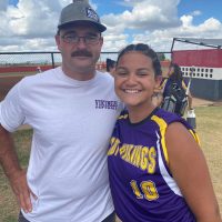 A smiling young softball player in a purple and gold jersey poses with an adult man in a grey baseball cap and white t-shirt at a sunny outdoor softball field.