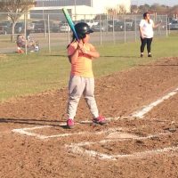 A young child in a batting helmet and orange shirt stands at home plate on a softball field, holding a bat.