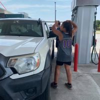 A young person wearing a grey jersey with the number 10 and black shorts is stretching by a white pickup truck at a gas station.