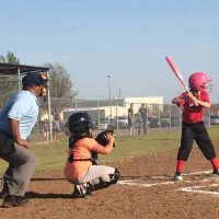 A young softball player with a pink helmet and red uniform is at bat, with a catcher and an umpire nearby