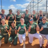 A group of eleven young softball players in green and white uniforms pose for a photo on a dirt field, making peace signs with their hands.