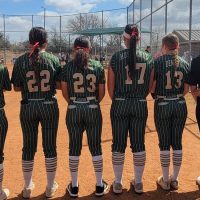 Ten softball players in green and white pinstripe uniforms stand with their backs to the camera on a dirt field, with a chain-link fence and trees in the background under a partly cloudy sky.