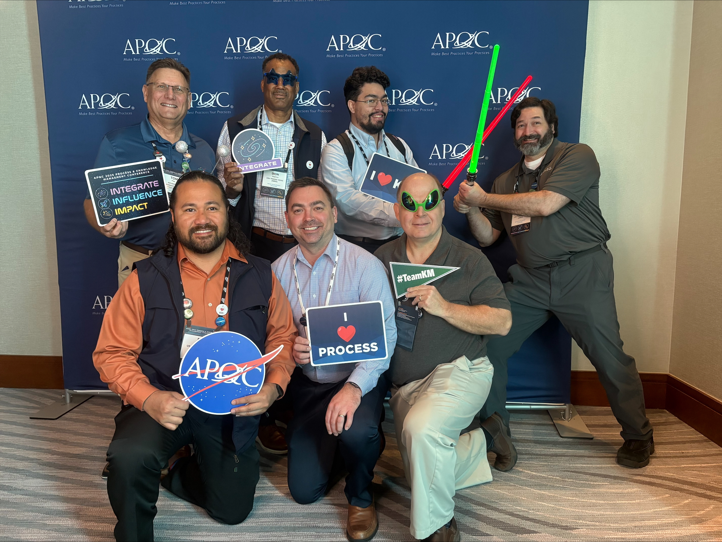 A group of eight individuals poses for a photo in front of a backdrop with the "APQC" logo. They are holding various signs and props, including a NASA logo, hearts with the words "INTEGRATE" and "PROCESS," and light sabers. Some are wearing sunglasses and name tags.