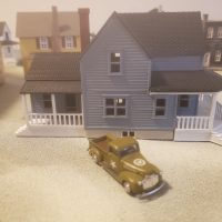 A miniature blue house with a white porch sits on a sandy surface, with a small olive green vintage pickup truck parked in front of it. Other miniature buildings are visible in the blurred background.