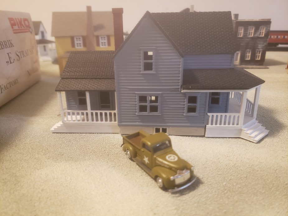A miniature blue house with a white porch sits on a sandy surface, with a small olive green vintage pickup truck parked in front of it. Other miniature buildings are visible in the blurred background.