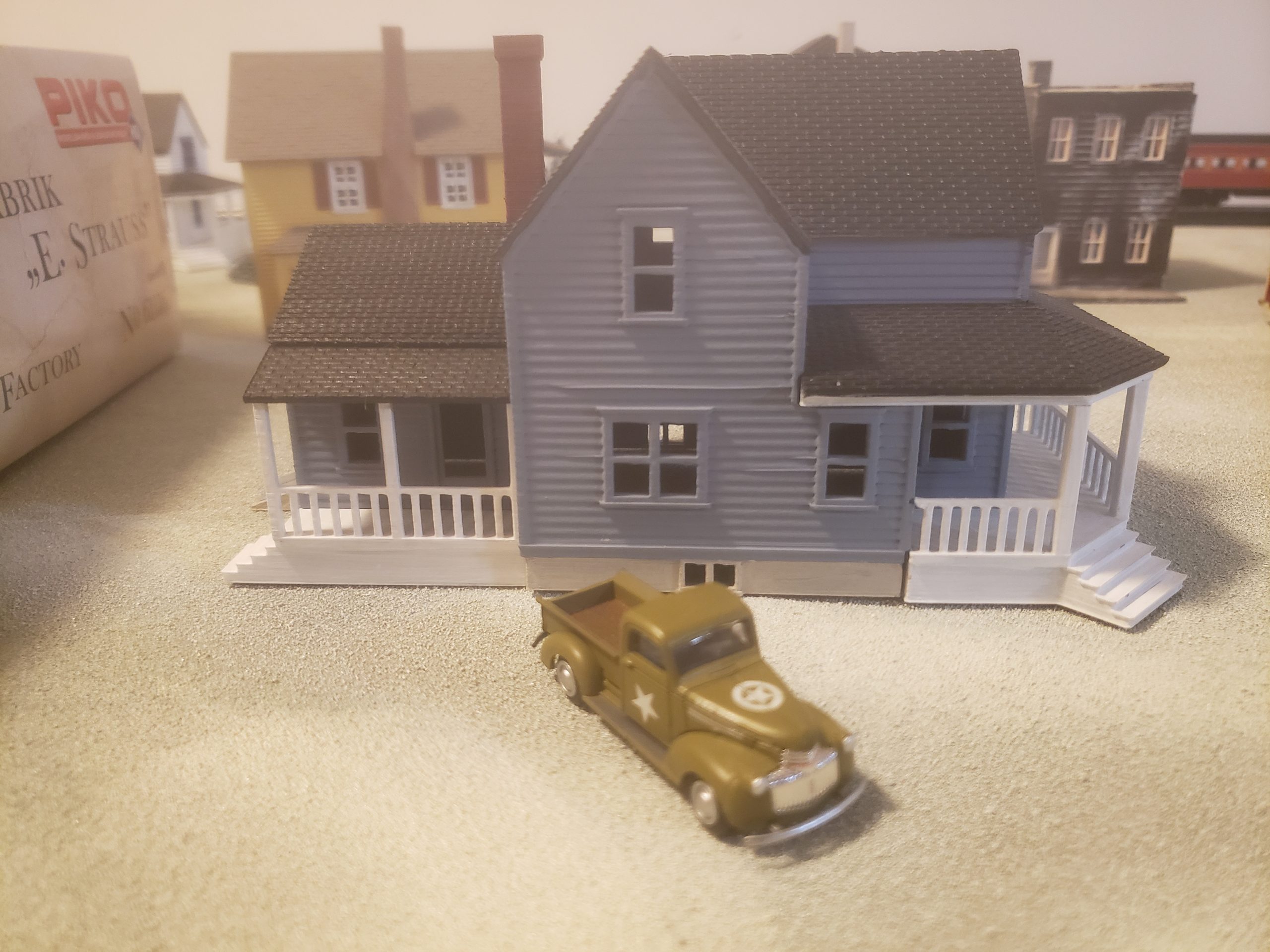 A miniature blue house with a white porch sits on a sandy surface, with a small olive green vintage pickup truck parked in front of it. Other miniature buildings are visible in the blurred background.