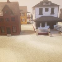 Two miniature houses, one red brick and one white with a porch, stand on a sandy surface, with other blurred miniature buildings in the background.