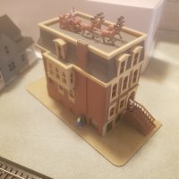 A miniature brown model building with Santa and reindeer on its roof. A small figure stands on the front steps, and another miniature house and train tracks are visible in the blurred background.