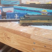 Two model trains, one green with "Maine Central" and one blue and white with "Boston and Maine," are positioned on a wooden surface with hobby boxes in the background.