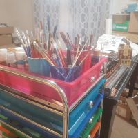 A close-up view of a pink, blue, and green rolling cart brimming with art supplies, including paintbrushes and paint bottles. In the background, a table holds model train sets and other hobby items.