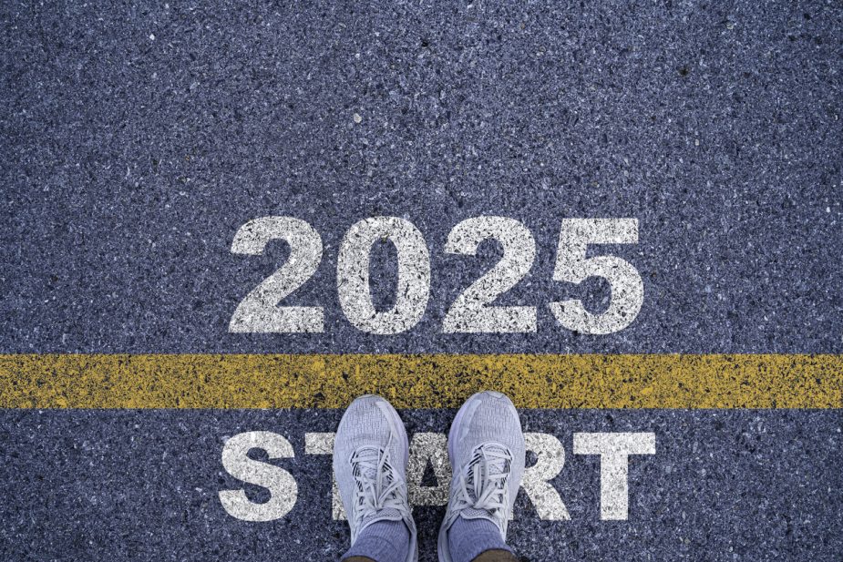 An overhead view of a person's feet in gray sneakers standing on asphalt. The numbers "2025" are painted above a yellow line, and the word "START" is painted below the line.