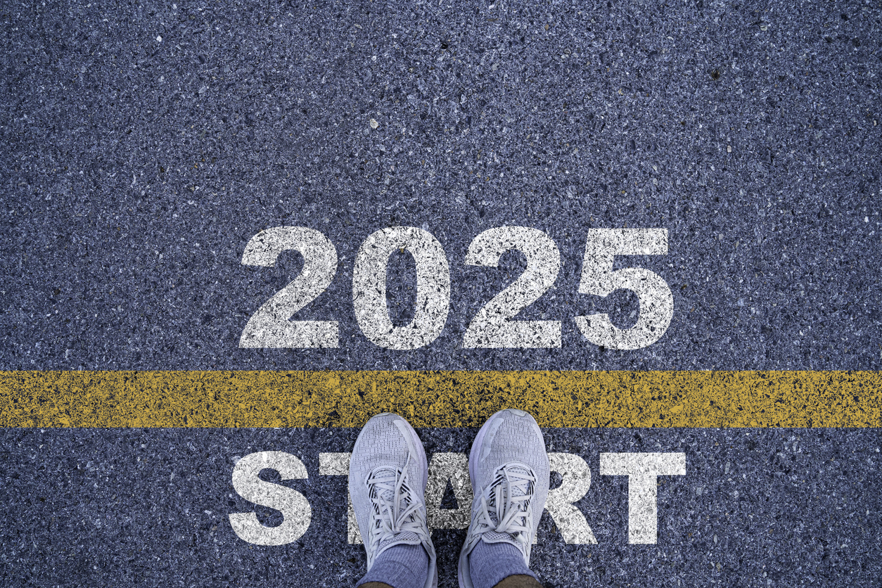 An overhead view of a person's feet in gray sneakers standing on asphalt. The numbers "2025" are painted above a yellow line, and the word "START" is painted below the line.