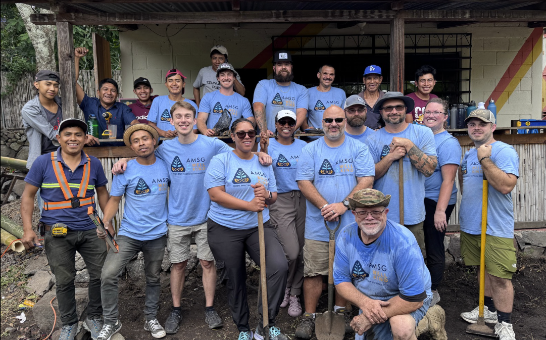 A large group of people, a mix of volunteers and locals, posing for a group photo outside a building, many holding tools like shovels and hammers. They are smiling and appear to be part of a humanitarian or service project.