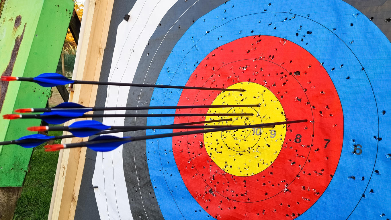 Several blue and red-fletched arrows are embedded in a red, yellow, and blue archery target, with multiple arrows hitting near the bullseye.