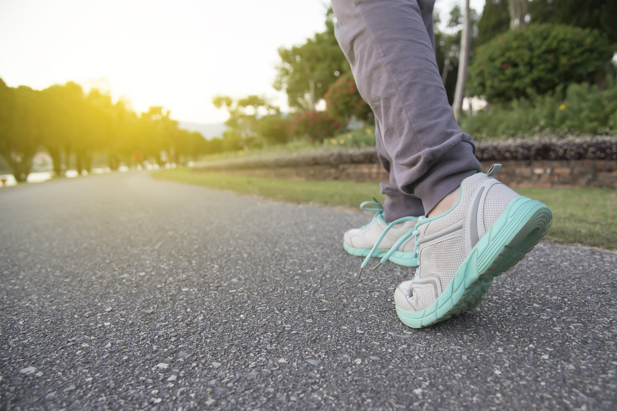 A low-angle, close-up shot of a person's legs and feet wearing light grey athletic pants and white and turquoise running or walking shoes, stepping forward on an asphalt path in a park with sunlight streaming from the left.