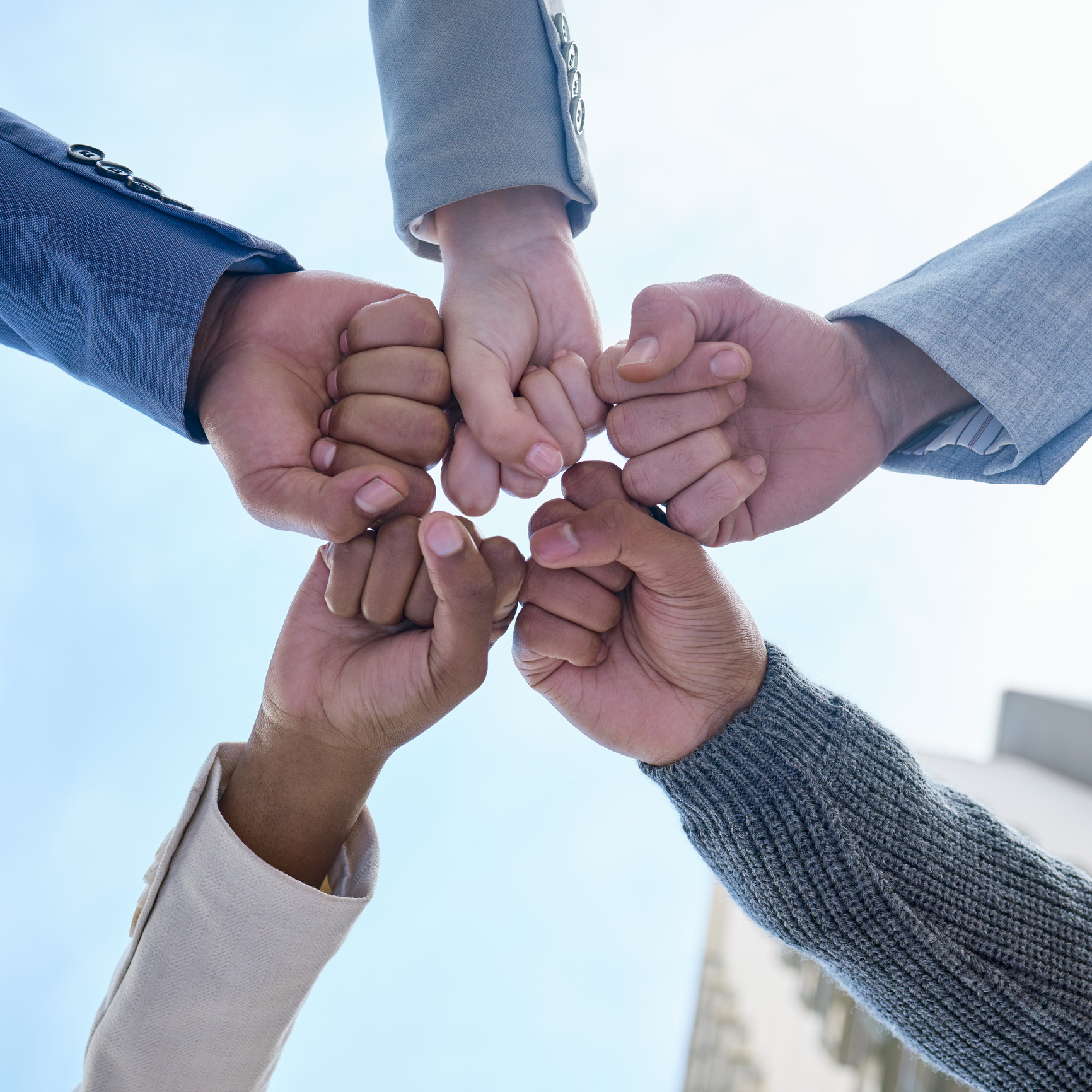 A low-angle shot of five diverse individuals in business attire, their fists meeting in the center to form a star shape against a bright blue sky, symbolizing teamwork, unity, and collaboration.