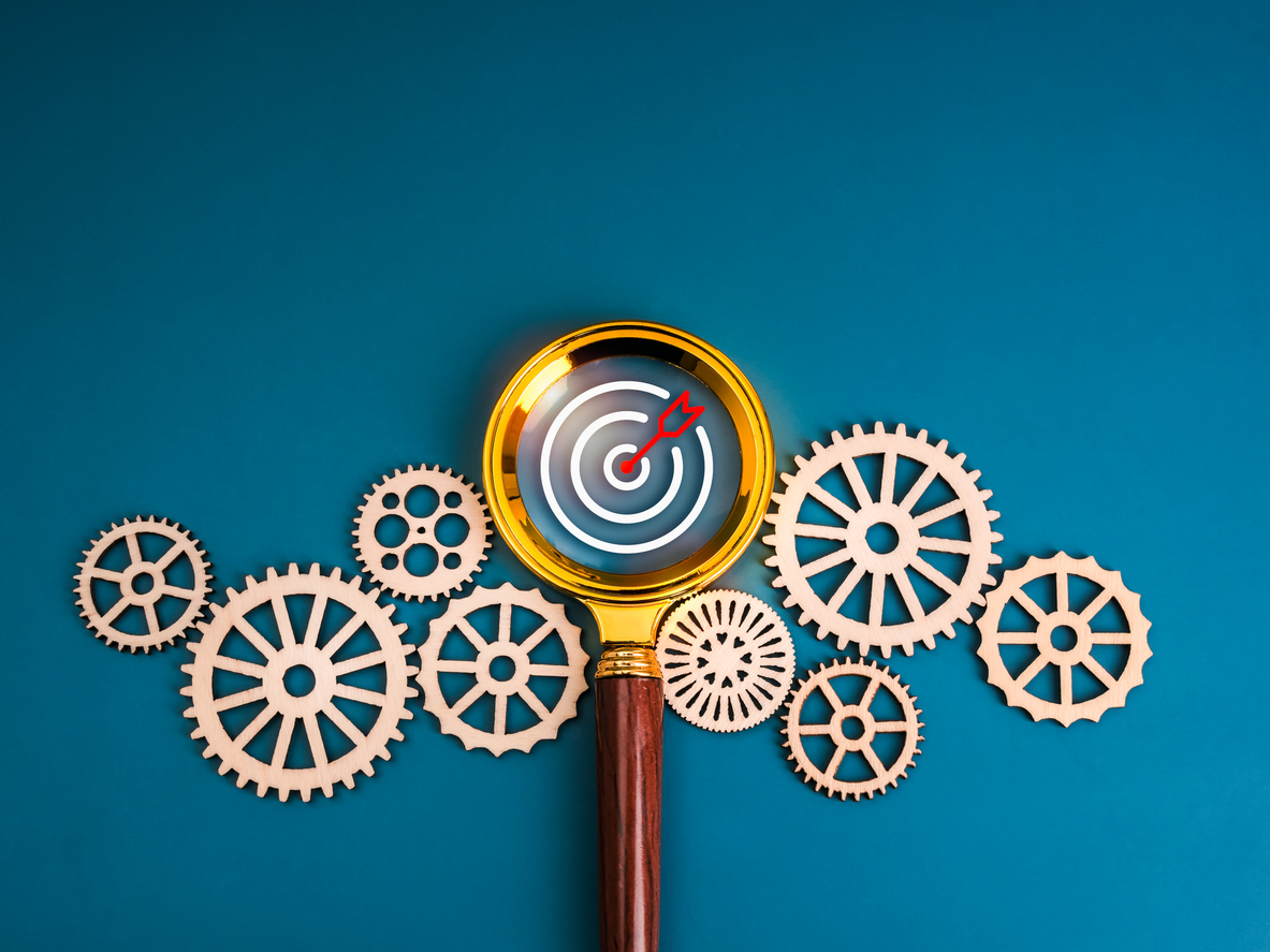 Magnifying glass focused on a target with an arrow hitting the bullseye, surrounded by a group of wooden gear cogs.
