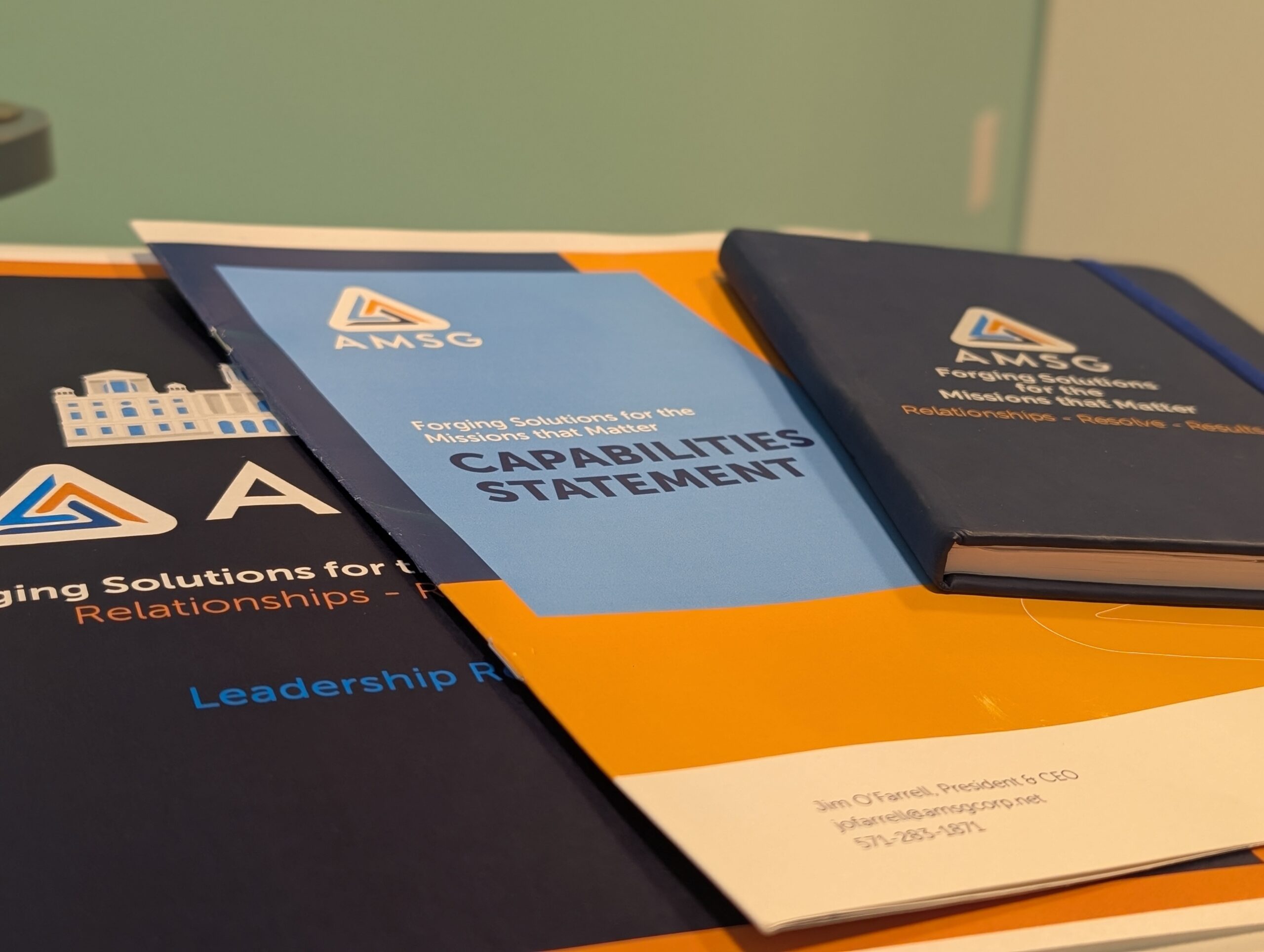 Stack of printed marketing materials and a dark blue notebook from AMSG. The blue and orange document on top is a 